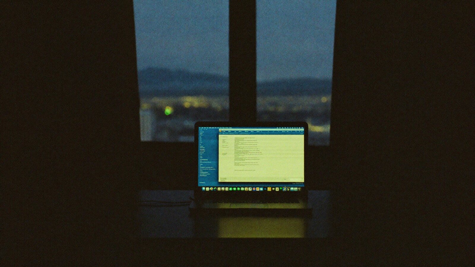 A laptop glowing on a desk at blue hour above a city skyline